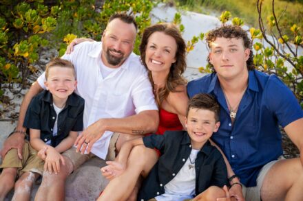 mom, dad and 3 children sitting closely together in sand and foliage on beach looking up and smiling