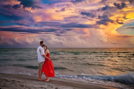women in red dress hugging man on Longboat Key beach with colorful cloud sunset backdrop