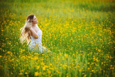 high school senior with long blond hair and white dress kneeling in yellow flower field fluffing hair