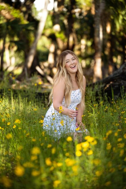 young woman with long blond hair sitting in wooded yellow flower field laughing