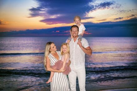 mom, dad, and 2 little children standing together smiling with colorful beach sunset backdrop