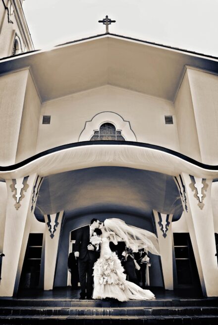 bride with veil flowing in wind, kissing groom on from steps of ornate church entrance with a cross at top of building