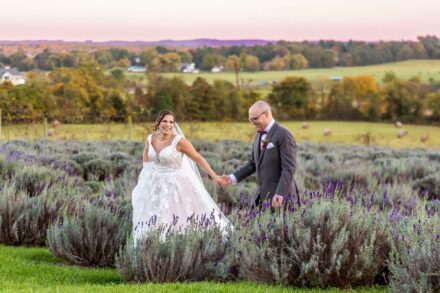 bride and groom smiling walking hand and hand through a lavender field with trees and rolling hills in background