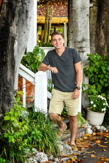 male high school senior in black v neck leaning again white wood stair rail in green setting