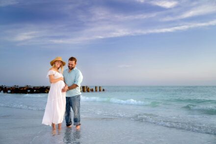 pregnant women in flowing white dress and wide brimmed hat standing on beach with husband touching belly