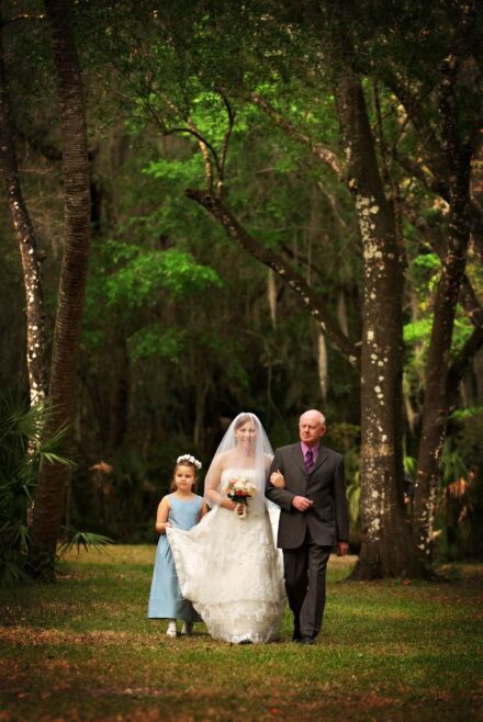 bride walking down forest tree line aisle arm in arm with dad and flower girl holding up her train