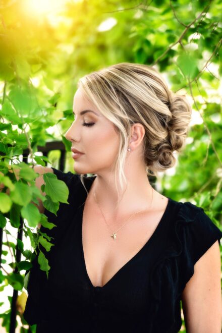 profile of blond woman in black top with sun coming through green trees in background
