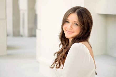 young woman with long brown hair sitting looking over shoulder smiling with white background