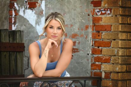 young blond girl leaning on metal railing with hand under chin with brick wall background