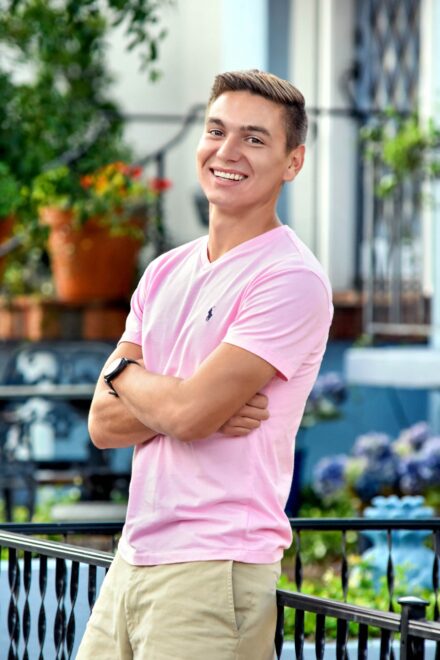 male high school senior with pink polo and arms crossed, smiling, leaning on railing