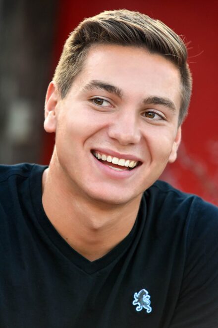 smiling headshot of male high school senior with brown hair, black polo, red background