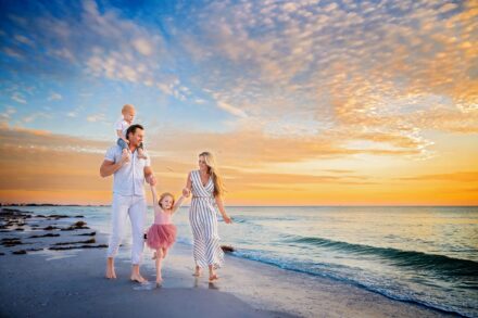 mom, dad and 2 little children walking along beach with sunset backdrop