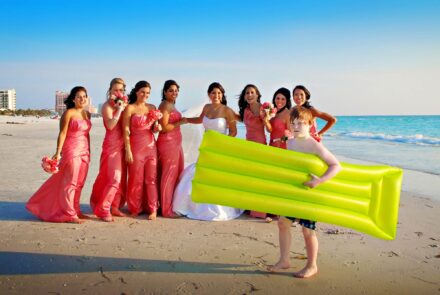 bridal party on beach laughing as young red headed boy unknowingly crosses in front with large neon green raft