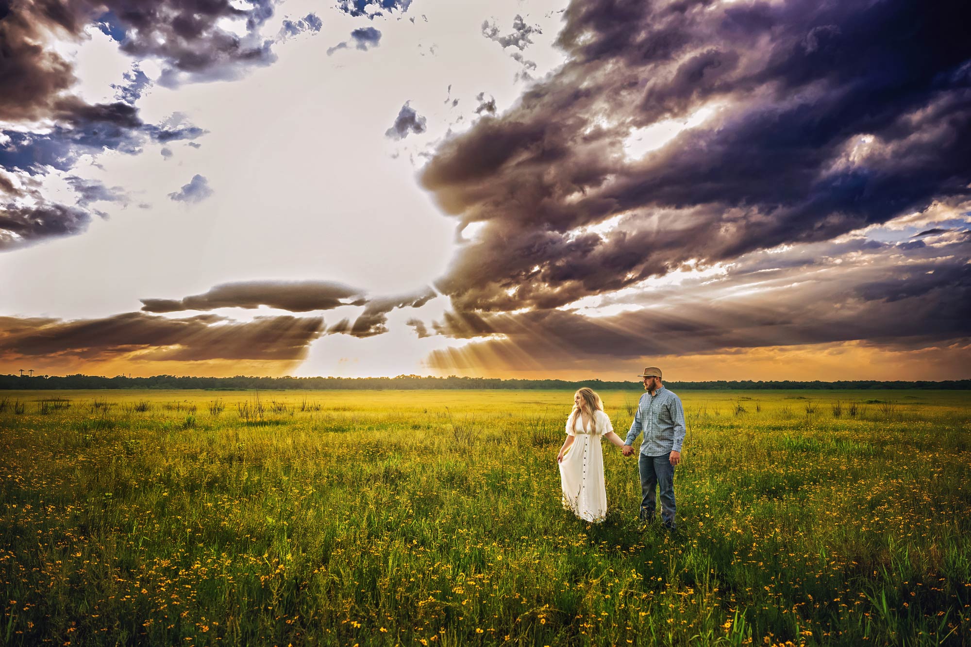 couple standing in yellow flower field holding hands with dramatic cloudy sky backdrop