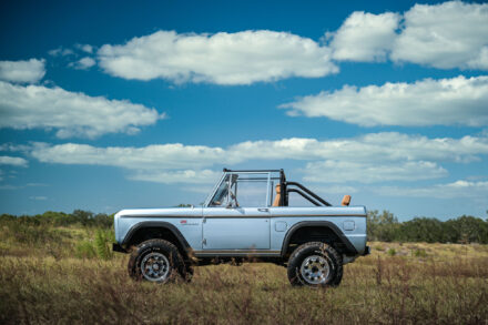 driver side view of vintage 1977 blue ford bronco sitting in a field with blue sky and white clouds above