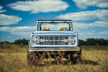 head on view of vintage 1977 blue ford bronco sitting in a field with blue sky and white clouds above