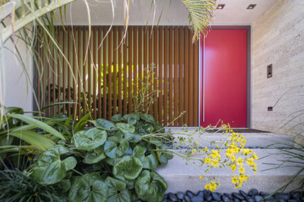 residential entrance with louver wall, red pivot door, green foliage and yellow flowers