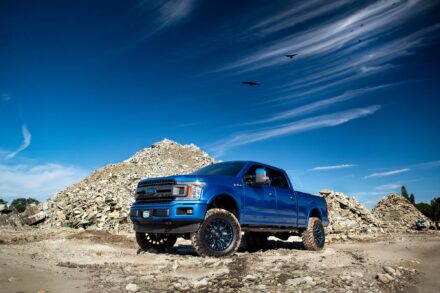 metallic blue ford f150 sitting in front of concrete rubble with blue sky and crows in air