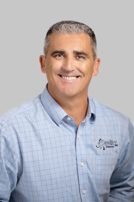 studio headshot of man with salt and pepper hair and blue button down shirt with quality enclosures logo on a light grey backdrop