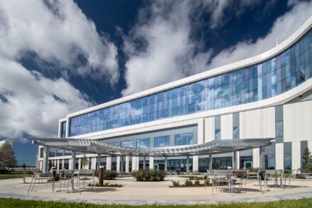 expansive white modern building with reflective blue window and an aluminum pergola feature above a sitting area with table and chairs