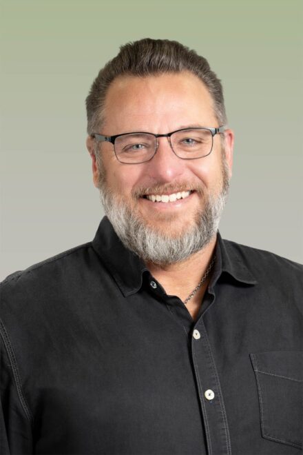 Professional studio headshot of man with glasses and beard on gradated green to gray backdrop