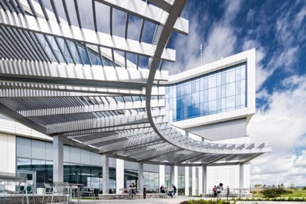 low wide angle of curved aluminum pergola structure with people sitting a tables below it, blue skies white clouds above