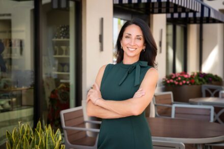 smiling brunette woman in green dress with arms crossed standing next to business windows