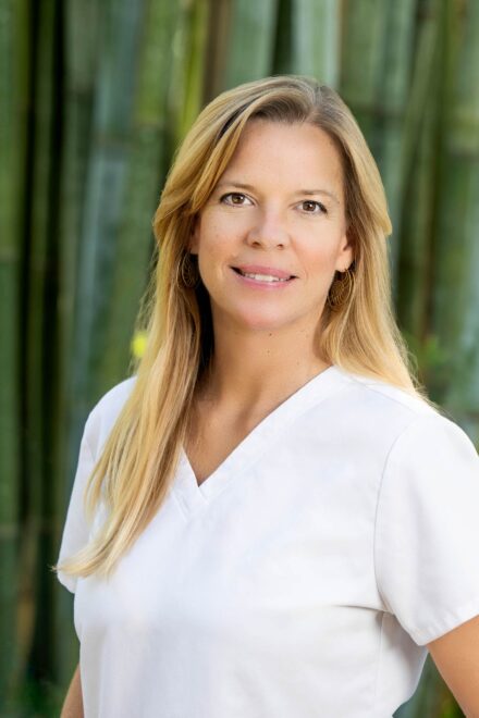 headshot portrait of a blond hair, brown eyed woman smiling wearing a white scrubs top standing in front of bamboo