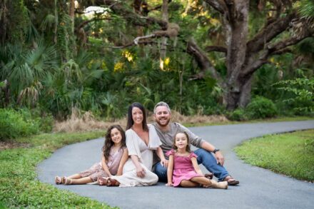 mother, father, and 2 daughters sitting together smiling in a natural setting with a moss covered oak