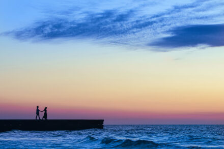 silhouetted couple walking holding hands on pier with beach waves and sunset sky