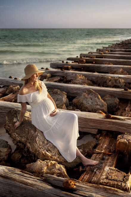 pregnant woman in long white dress and hat sitting holding belly on rocky pier at bradenton coquino beach