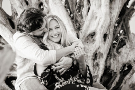 woman smiling while being hugged by a man sitting in front of weathered beach tree
