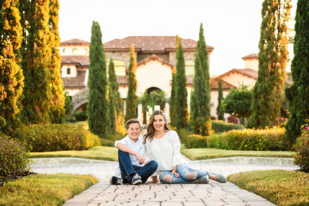 sister and brother sitting on pavers posing and smiling in front of well-landscaped large building with clay tile roof