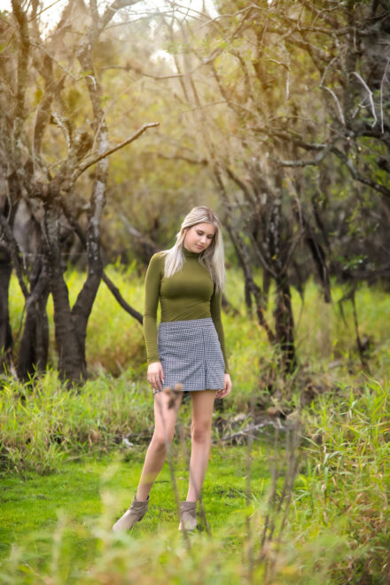 high school senior in black and white checkered skirt and long sleeve green top standing in green wooded area