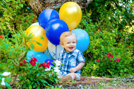 a blond baby boy sitting in a flower garden setting smiling, with blue, yellow, and white balloons behind him