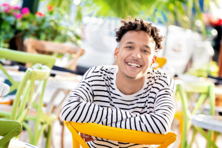 male high school senior with. black and white striped shirt smiling sitting backward on a yellow chair in an outdoor cafe setting