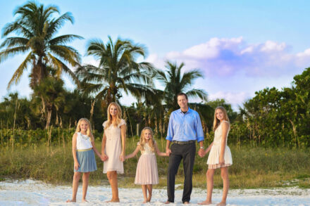 mother, father and 3 daughters holding hands and smiling in front of palm trees and sea oats on siesta key beach