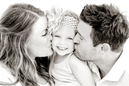mother and father kissing their smiling daughter with big floral headband on both cheeks