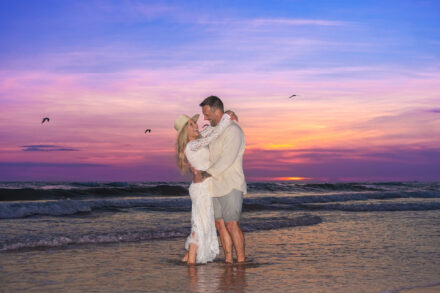 woman in hat and white dress smiling and holding man smiling back on beach at sunset