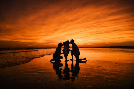 silhouette of mom and dad kneeling and kissing daughter and son on beach with orange cloudy sunset background