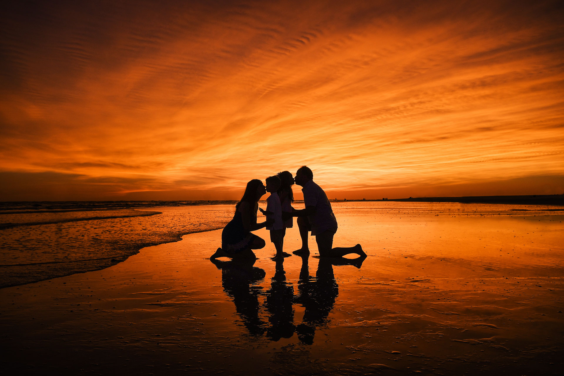 silhouette of mom and dad kneeling and kissing daughter and son on beach with orange cloudy sunset background