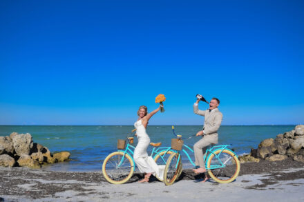 smiling bride and groom on bikes with the sarasota bay beach and rocks behind them