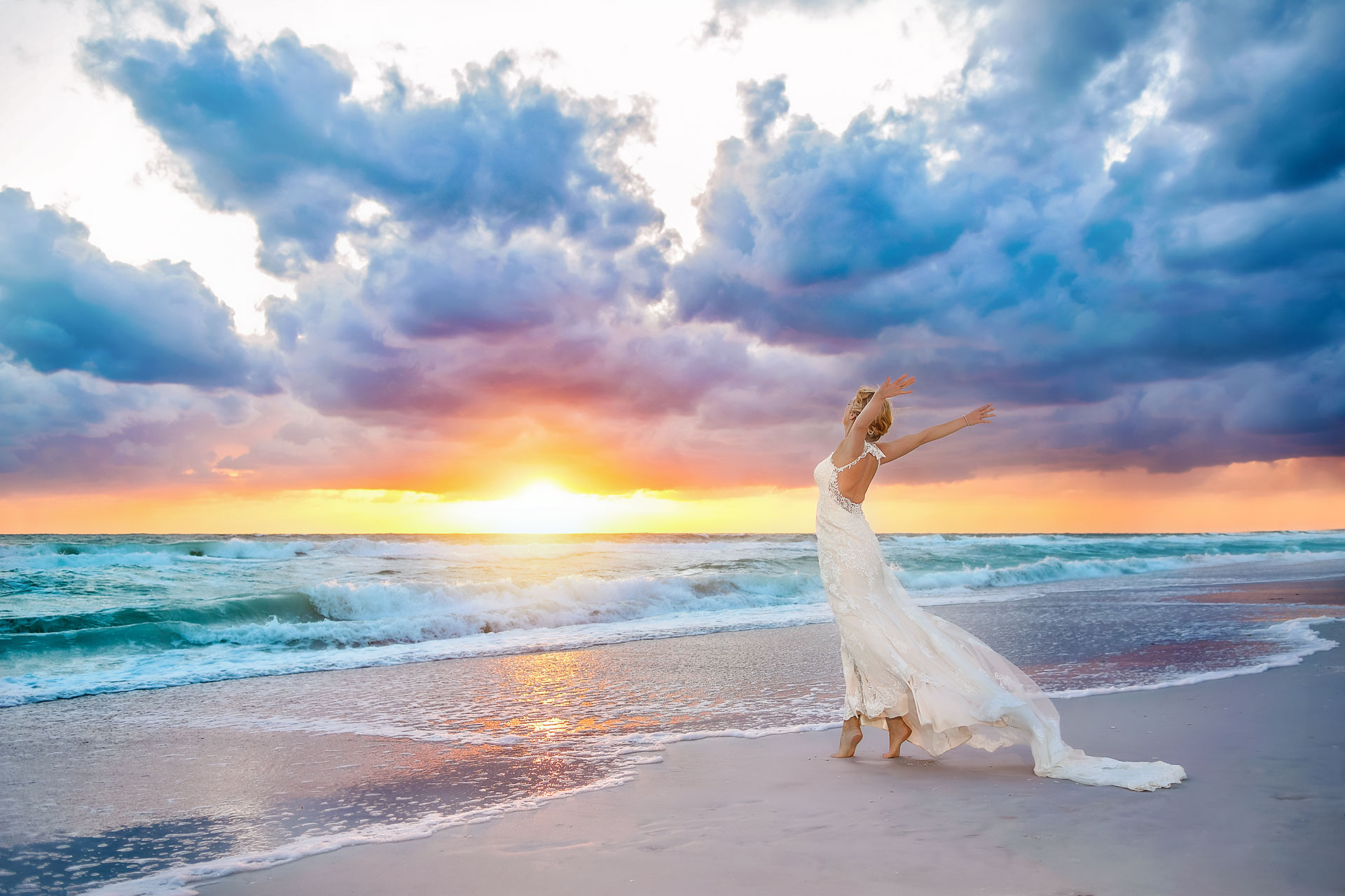 bride in wedding gown standing on tiptoes on beach with arms in the air facing a colorful cloudy sunset