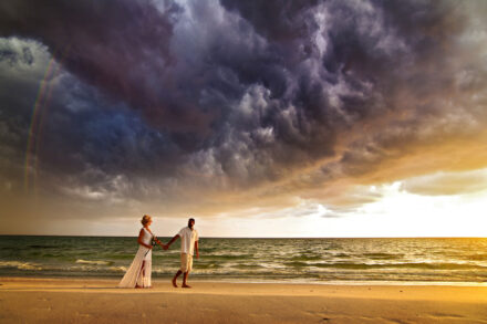bride and groom walking hand in hand on beach with dark cloudy sky
