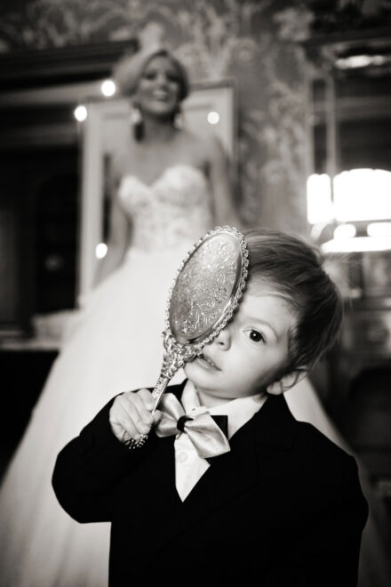 ring bearer holding mirror to face with bride in background laughing